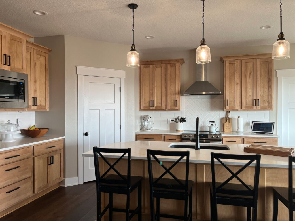 A kitchen with an island, light brown cabinetry, white countertops, and black accents and chairs.